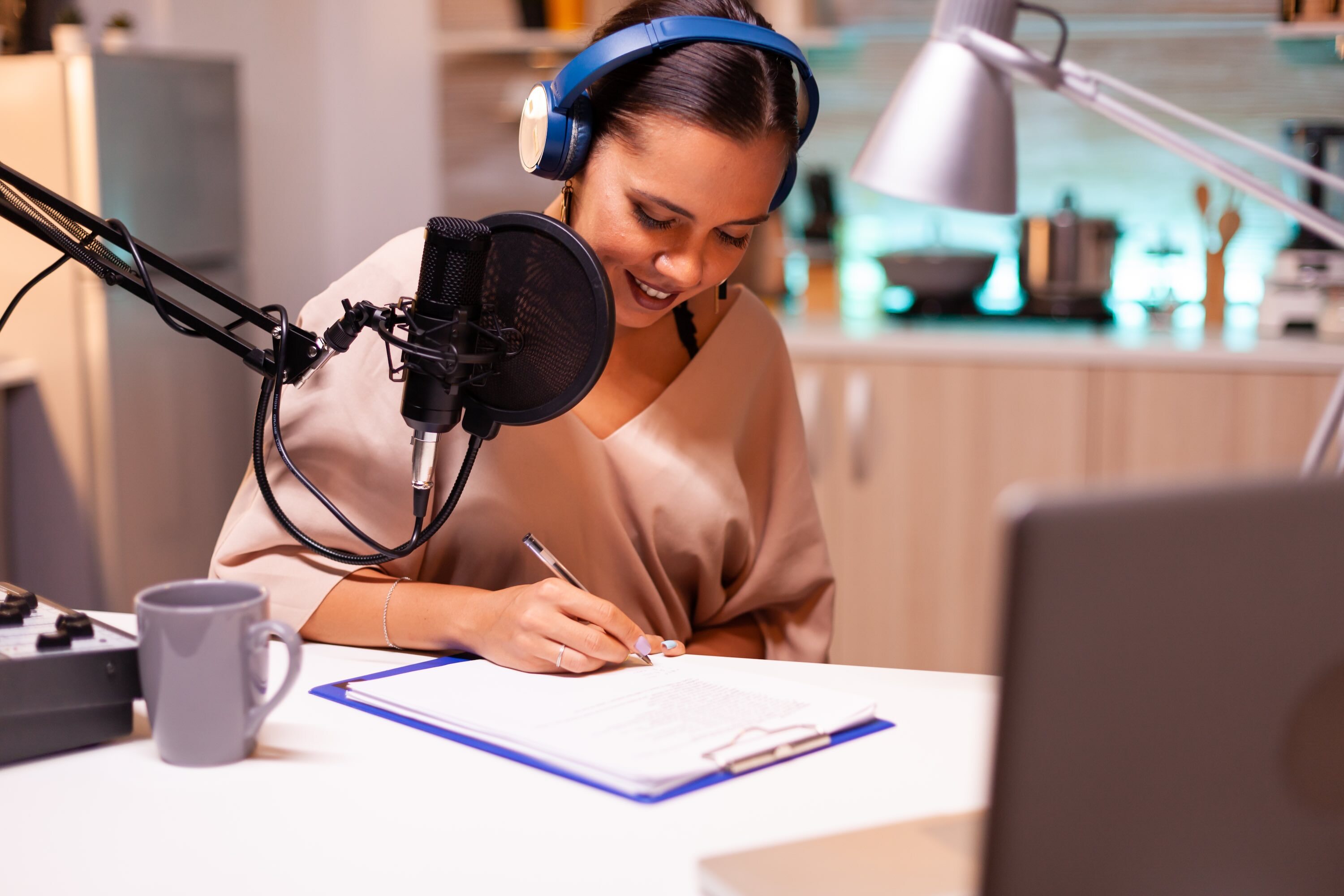 A woman wearing blue over-ear headphones writes notes on a clipboard at a desk, speaking into a studio microphone, with a laptop and coffee mug nearby in a home kitchen setting.