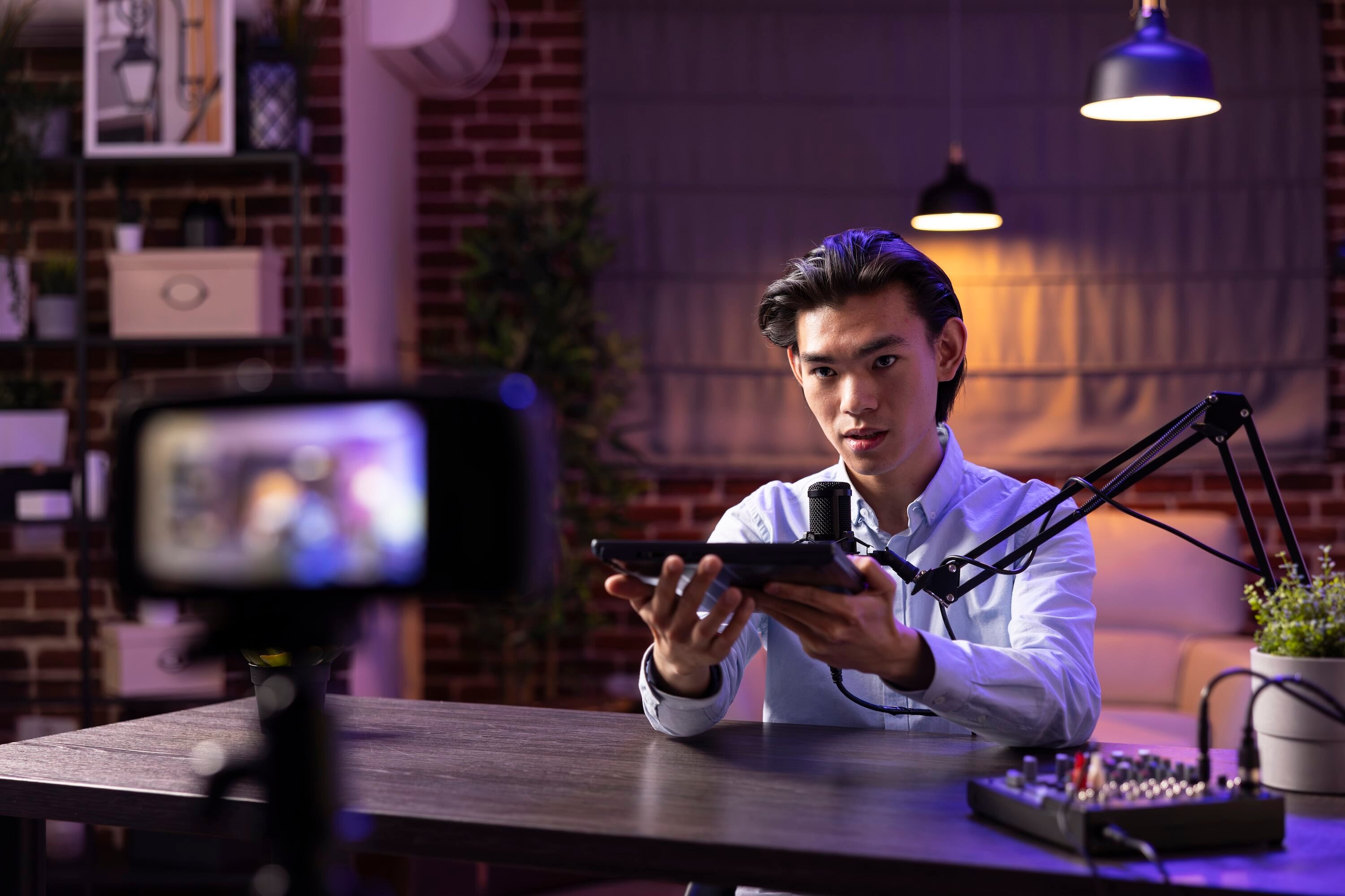 A man sits at a desk in a studio-like room, speaking into a microphone while holding a tablet, with a camera filming him and audio equipment on the table under warm and purple lighting.