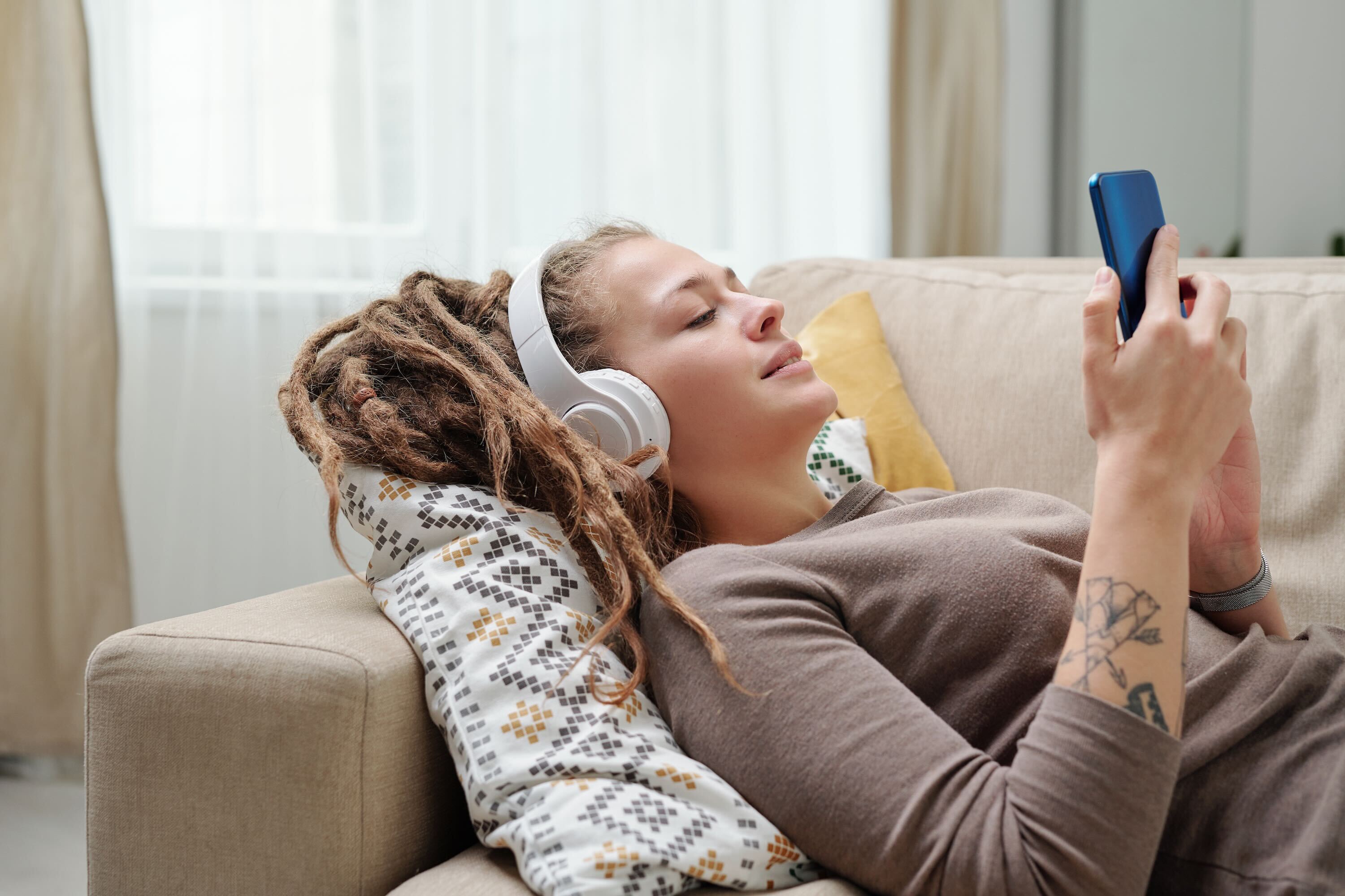A young woman with long dreadlocks lies on a beige couch, wearing white over-ear headphones and scrolling on a smartphone, her head resting on a patterned pillow in a bright living room.