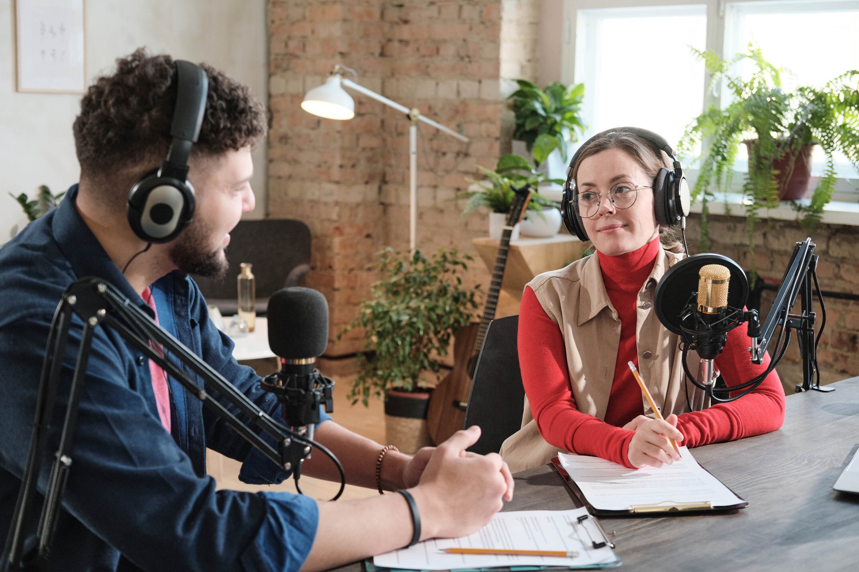 female sitting on desk wearing prescription glasses and brunette hair, with headphones, microphone equipment and taking notes. On her left, a make, with brown curly hair, headphoens and also microphone equipment, is speaking to her.