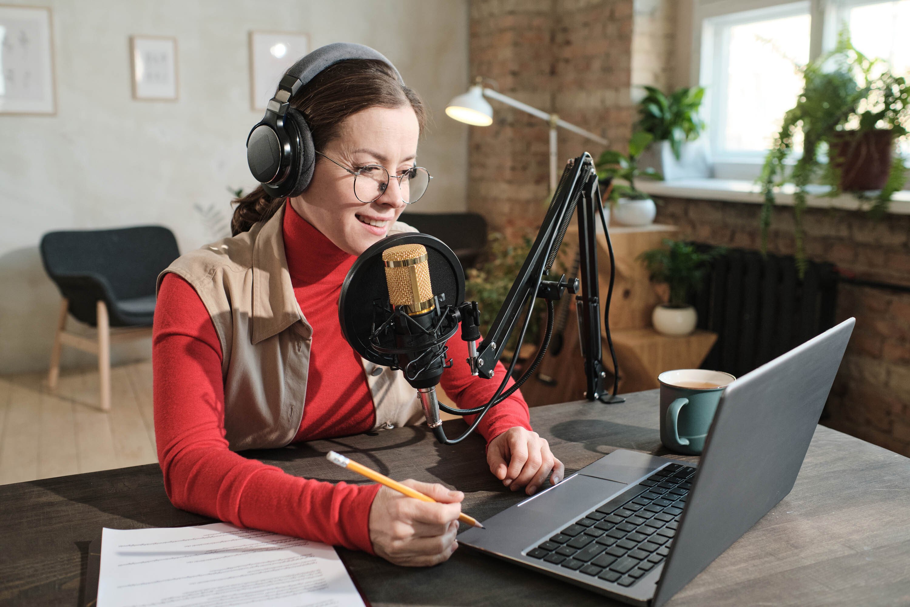 female sitting on desk wearing prescription glasses and brunette hair, with headphones, microphone equipment and laptop.