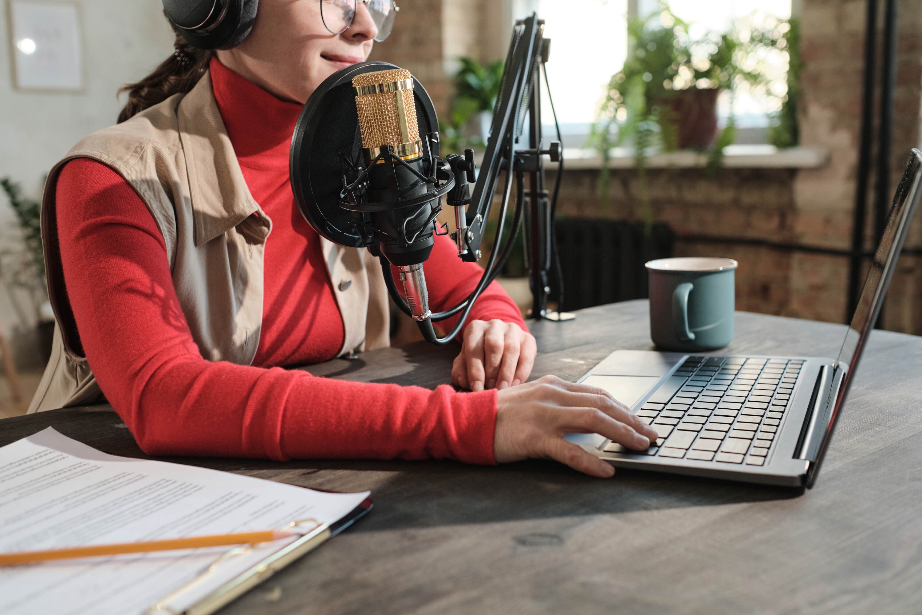 female sitting on desk wearing prescription glasses and brunette hair, with headphones, microphone equipment and laptop.